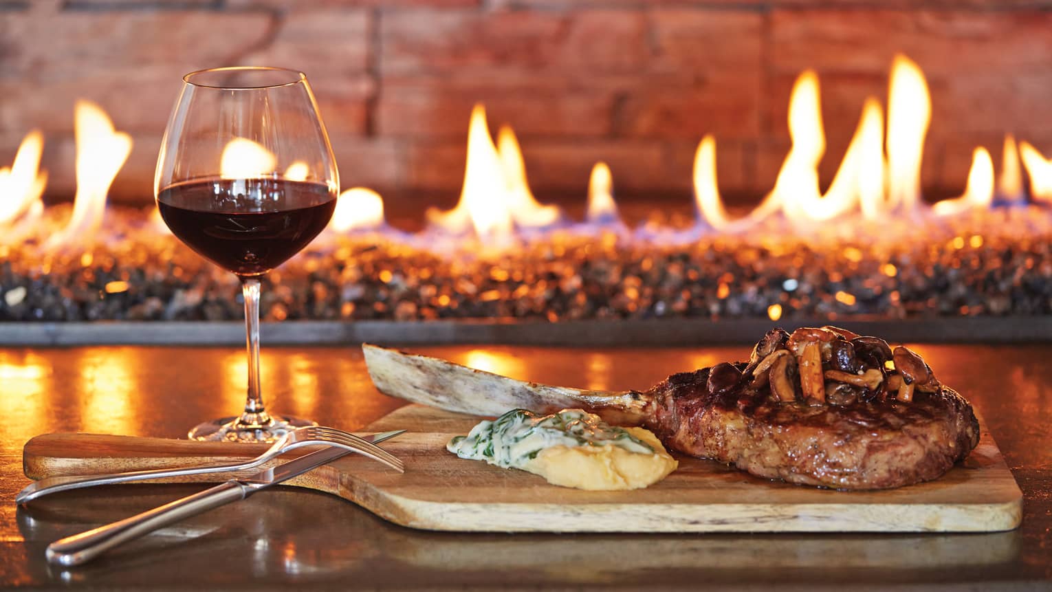 Glass of red wine, grilled chop on wood platter with cheese, mushrooms by fireplace