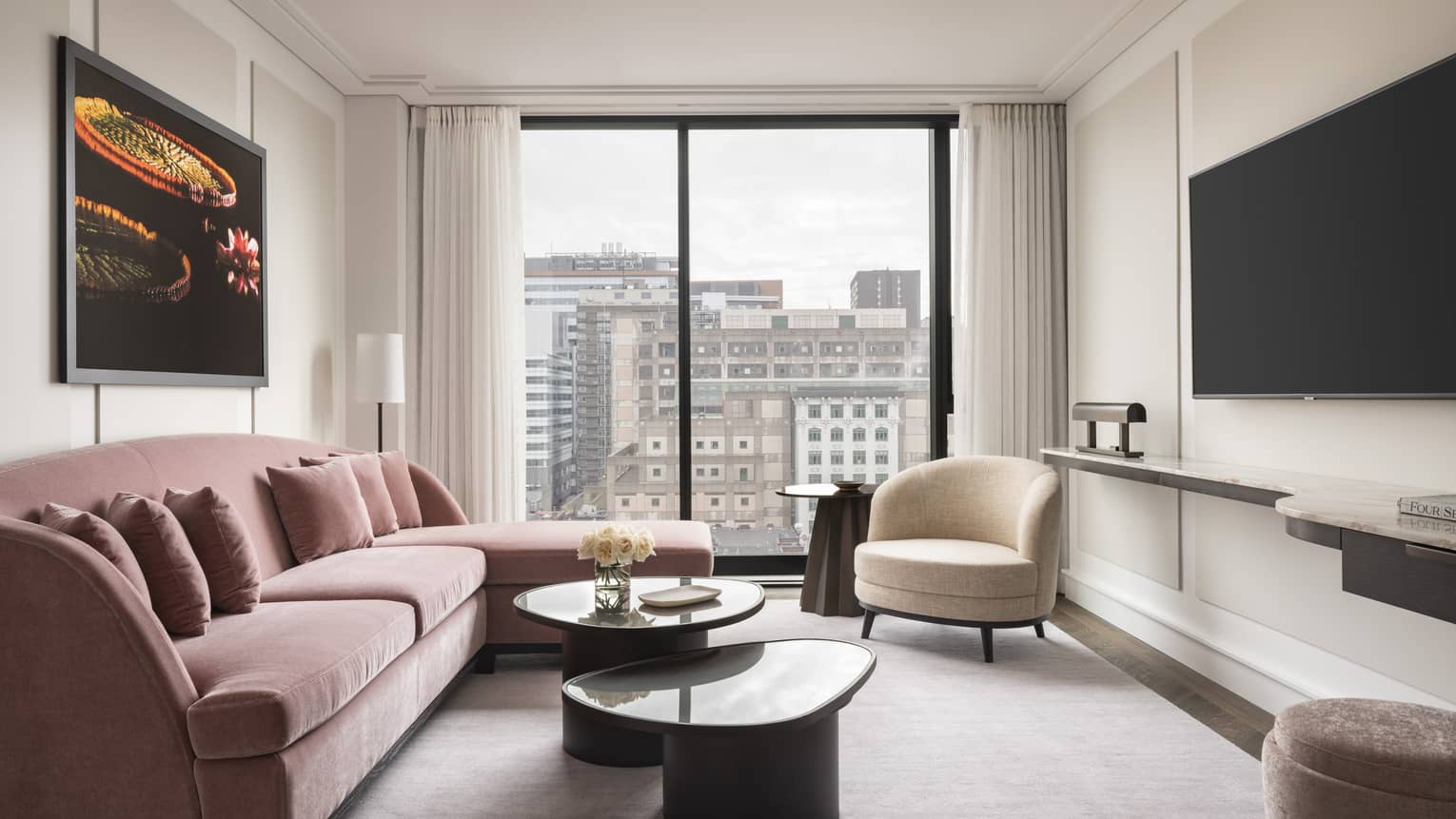 A seating area featuring a pink L-shaped couch, a circular white loveseat, and an cityscape view from a full-length window.