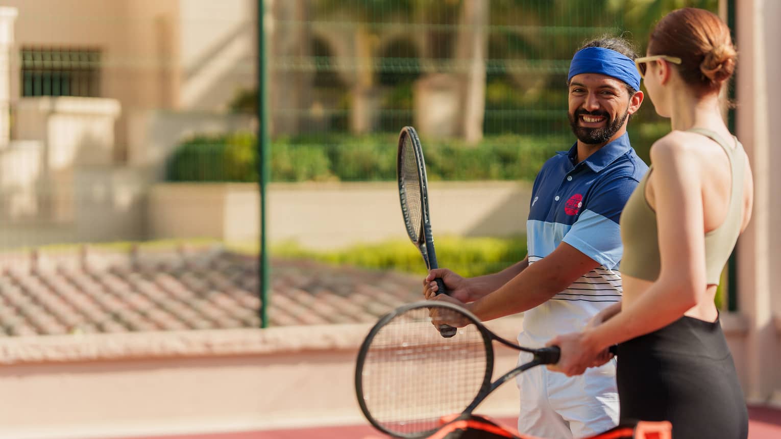 Two guests in athletic wear hold racquets on an outdoor court, one demonstrating a grip to the other, smiling.
