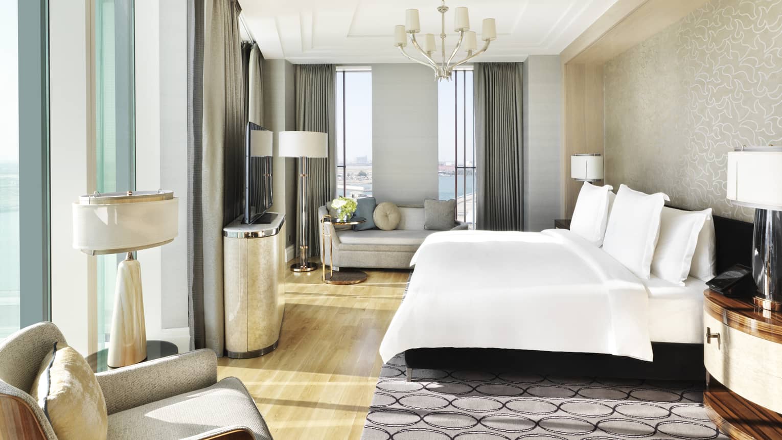 Executive Suite bed with white linens under chandelier, sunny corner windows, grey loveseat sofa and chair