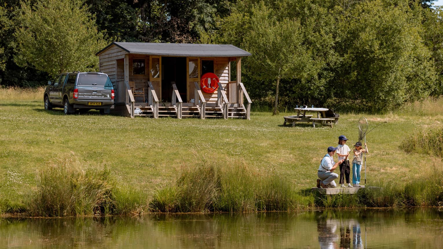 Three people fishing on small platform by lake with cabin-like building and picnic table in backdrop