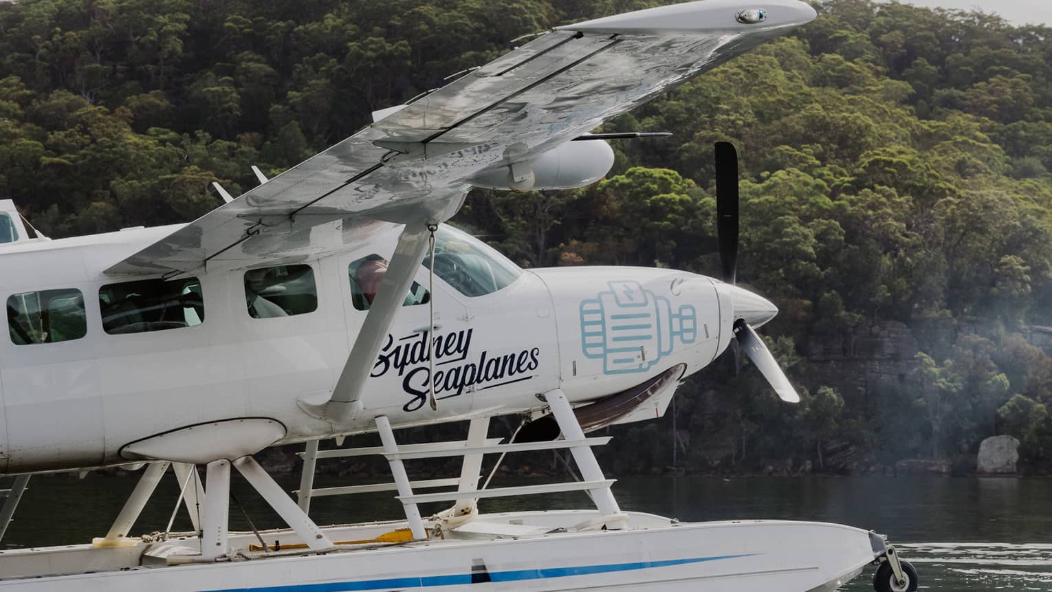 A white seaplane with the words "Sydney Seaplanes" in blue on its side, sitting on water with a lush treed hill behind it.