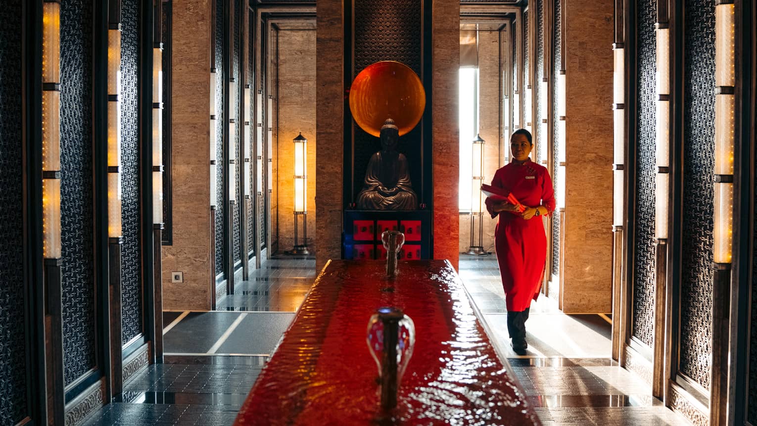 Person dressed in red walking down long hallway with fountain and Buddha statue