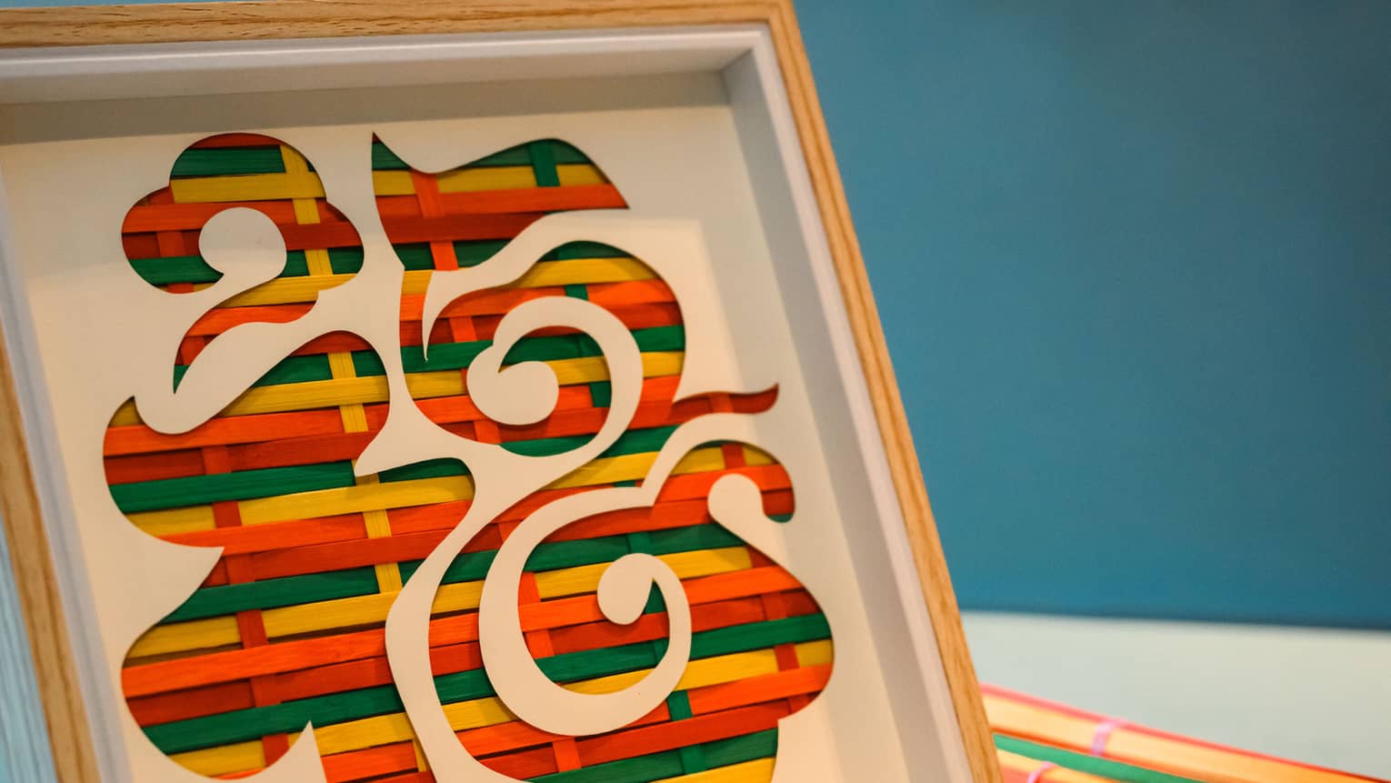 Coloured strips of straw woven together in wooden frames, creating an art pattern by itself or placed behind a stencil.