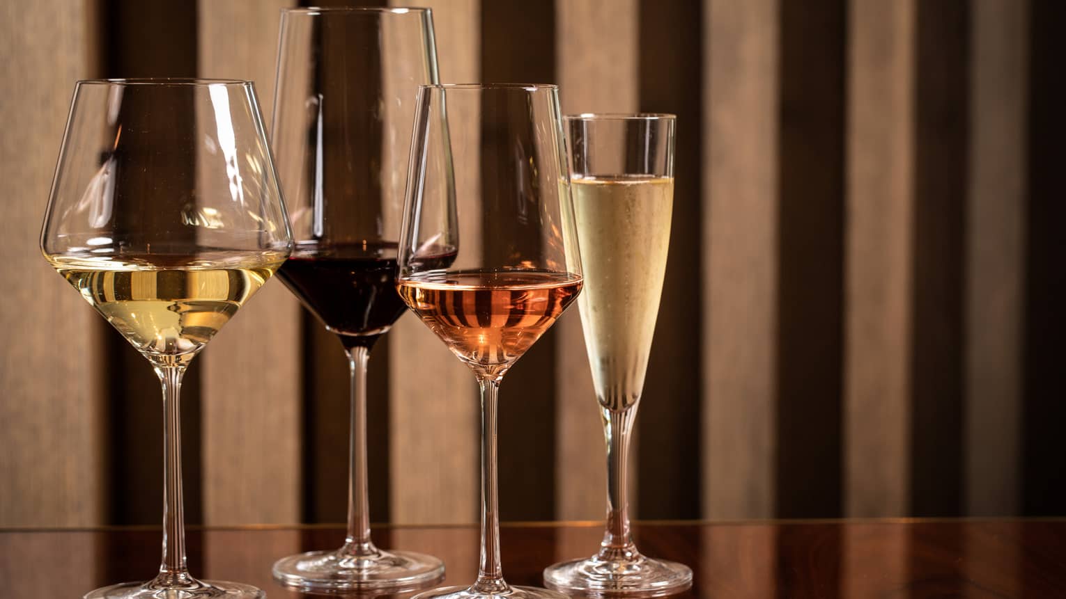 Four modern wine glasses holding white, red and rose wine are lined up on a wooden table 