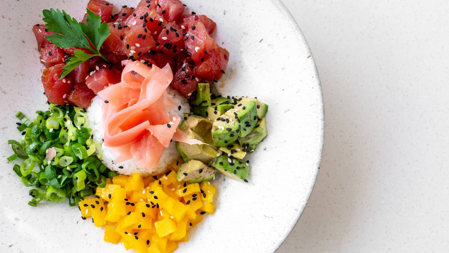 Poke bowl with chunks of tuna, mango, scallions and avocado plated with a rice, cilantro, sesame seeds and orange ponzu