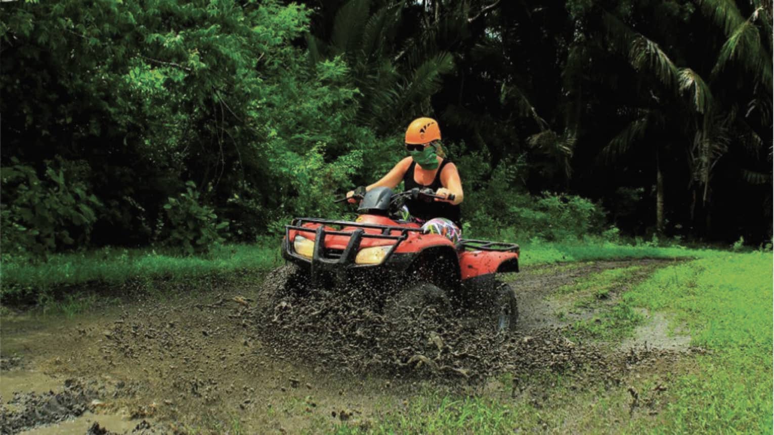 Person wearing a black tank top and bright orange helmet rides through the mud on a red ATV