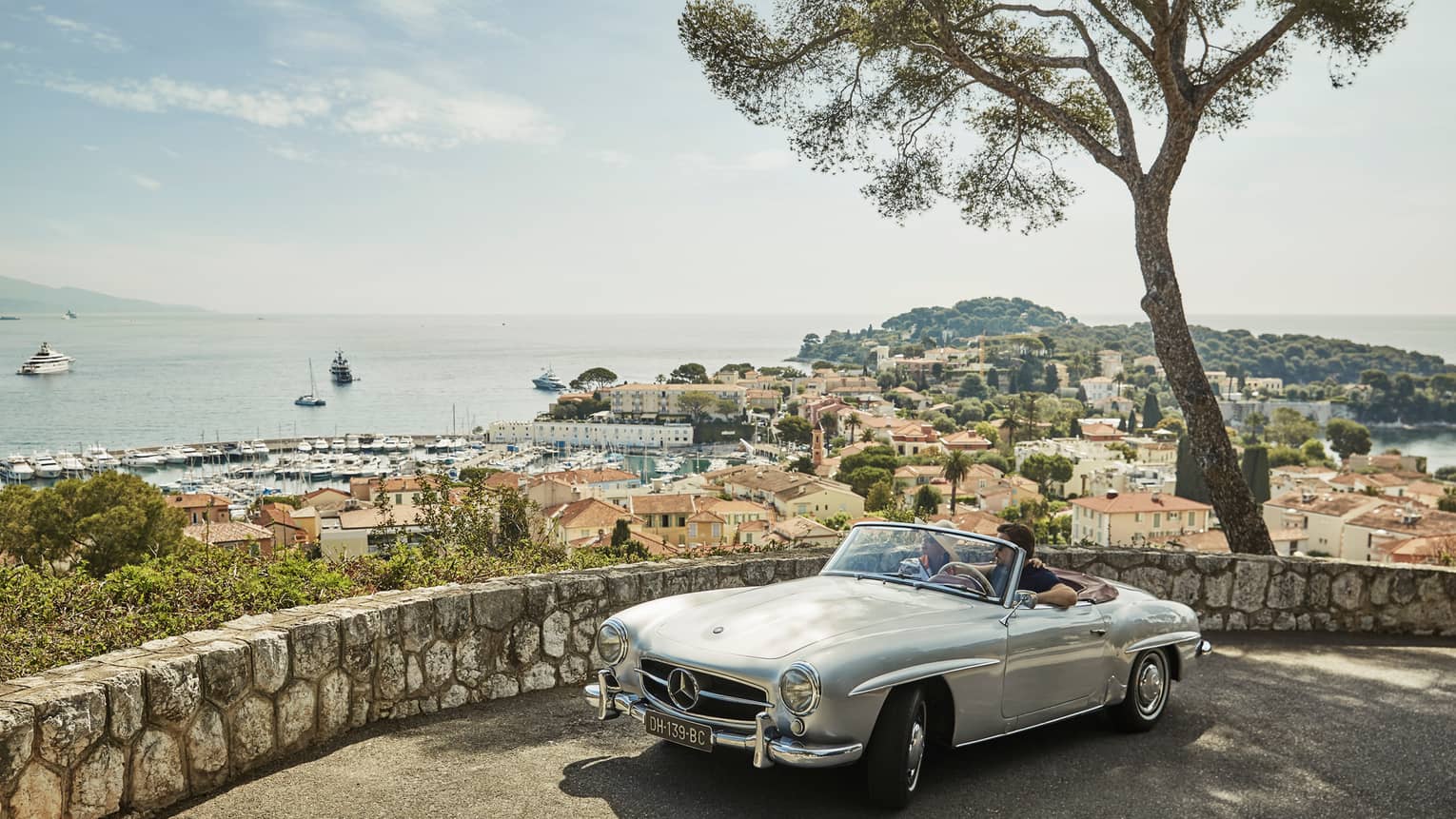 Couple in luxury vintage white car parked under tree by stone wall overlooking red roofs, water