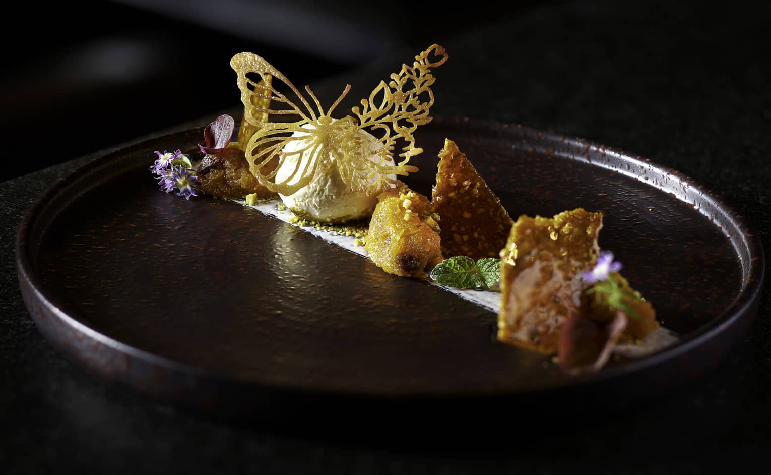 Traditional Indian dessert with butterfly-shaped tuile, edible flowers and fresh mint on black earthenware plate