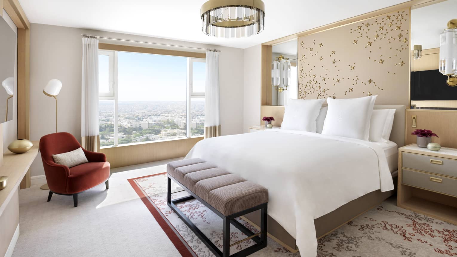 A large floral-themed bedroom with a bed in the centre, and a burgundy chaise beside a large bay window overlooking the city. 