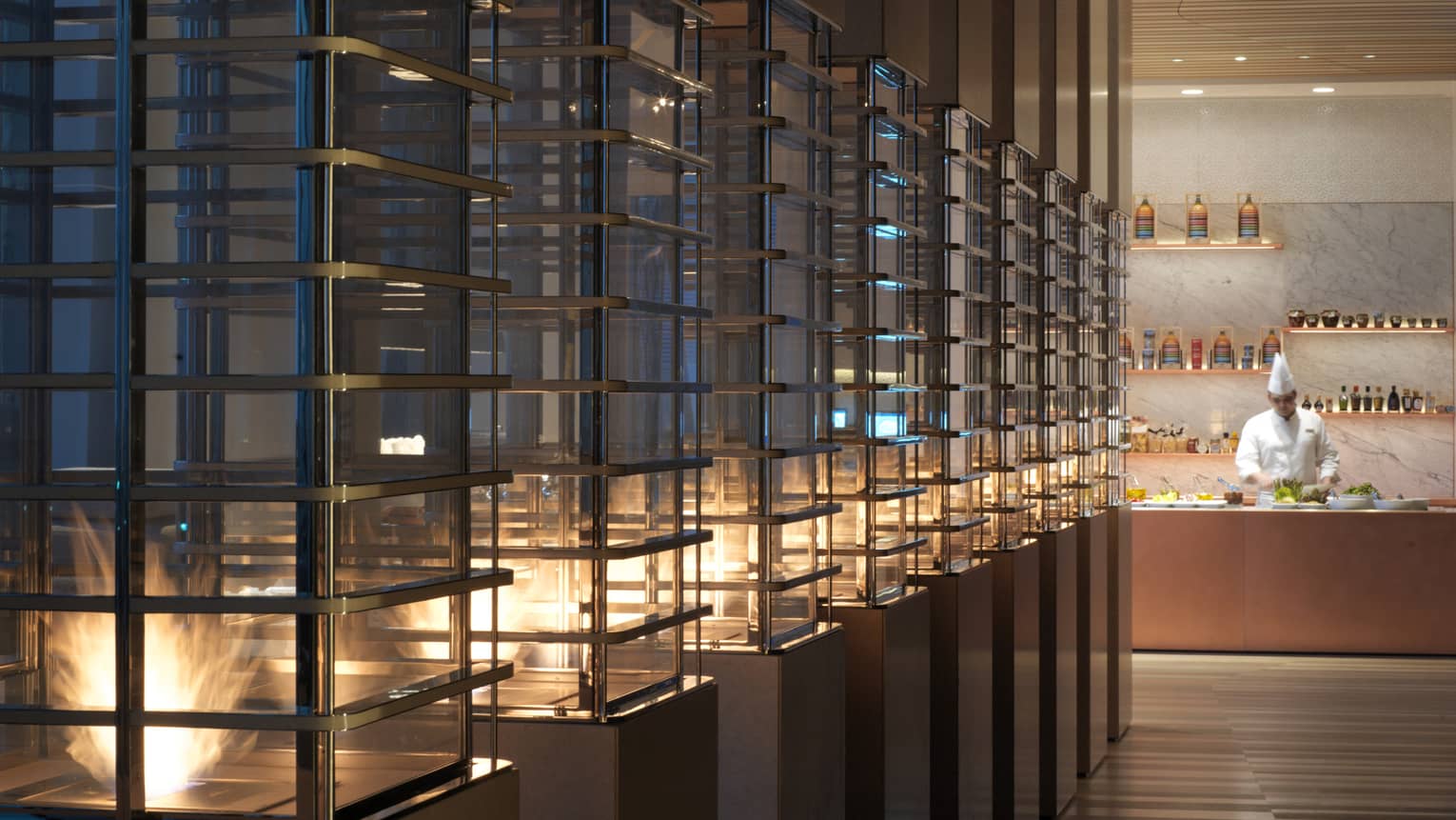 Row of pillars with glass, flames in dimly-lit room, counter and chef in background