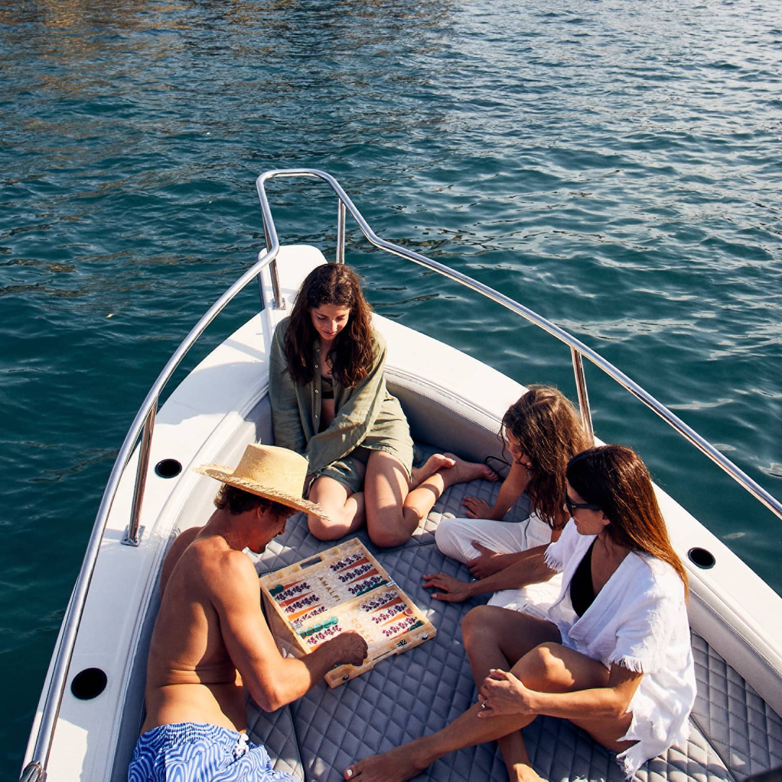 People relax and play a board game on a boat while floating on blue-green waters, with the rocky, hilly coastline in the background.