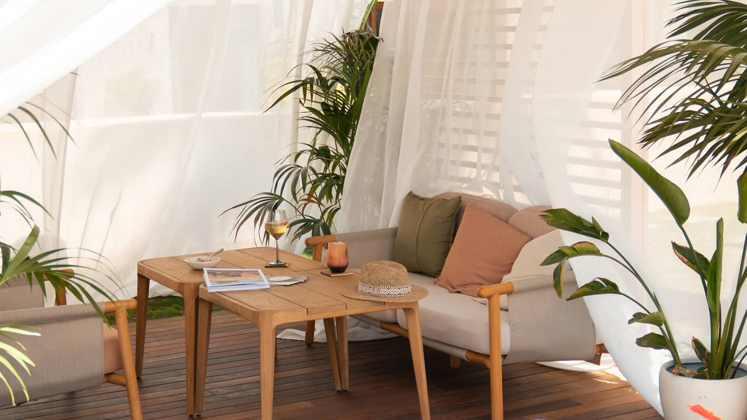 Inside of cabana with wooden floors, sheer white curtains, sofa, coffee table and tropical plants