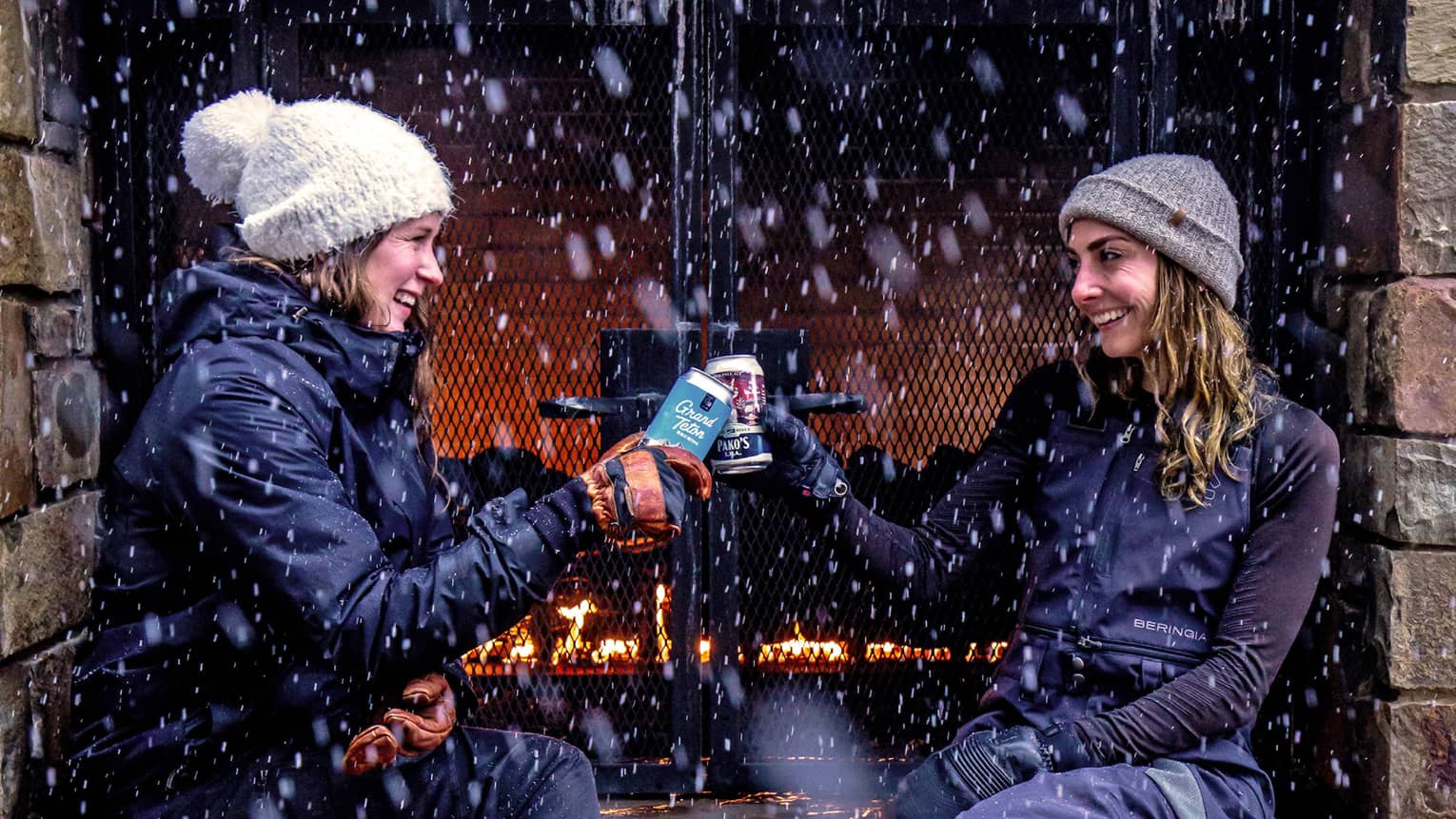 Two women drinking outside near a fire while it is snowing.