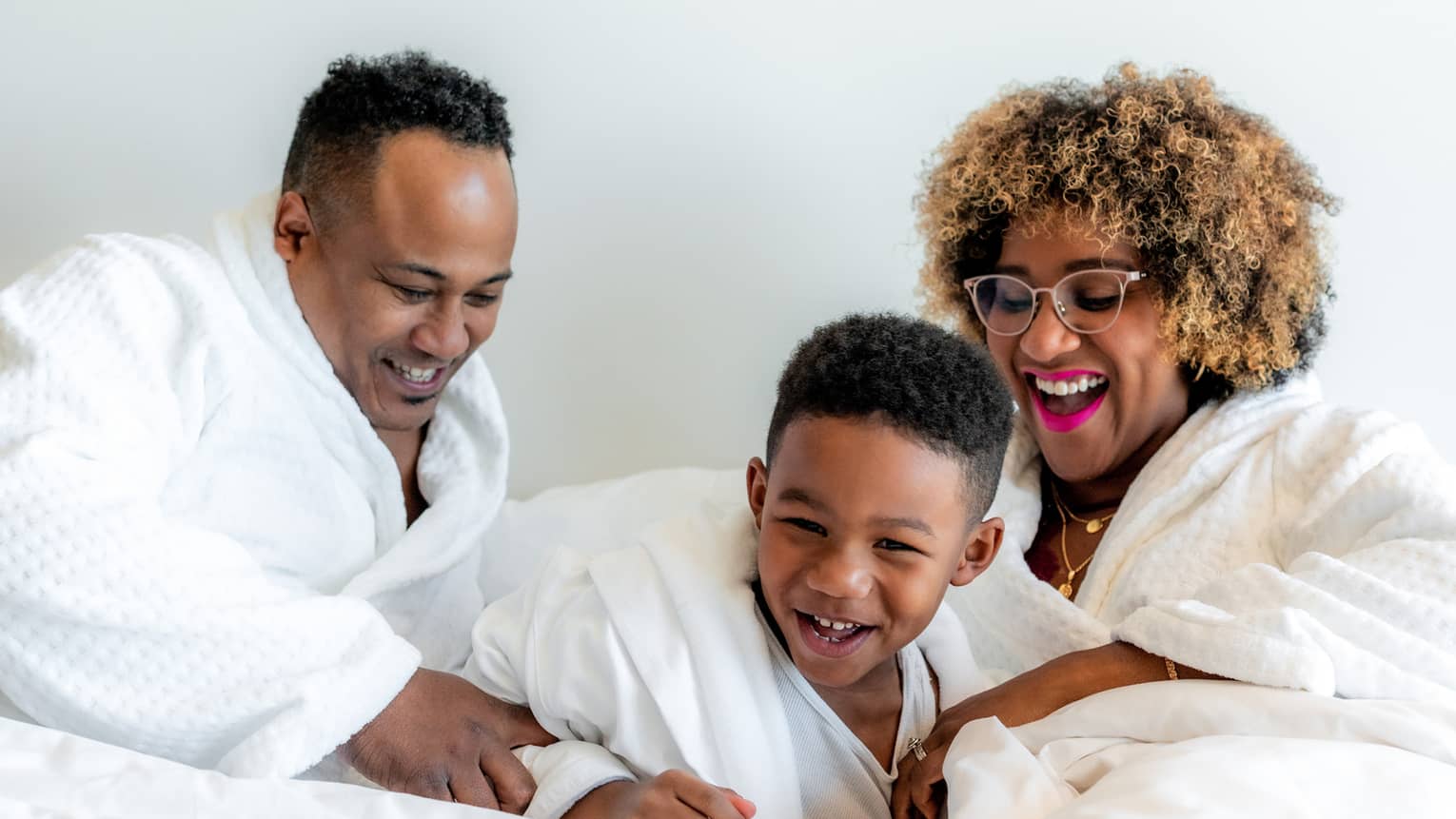 A mother, father and son playing on a white bed.