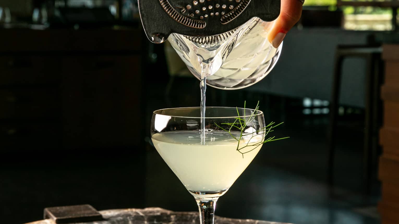 A hand pours a cloudy yellow liquid through a metal cocktail strainer into a martini glass garnished with fennel fronds.