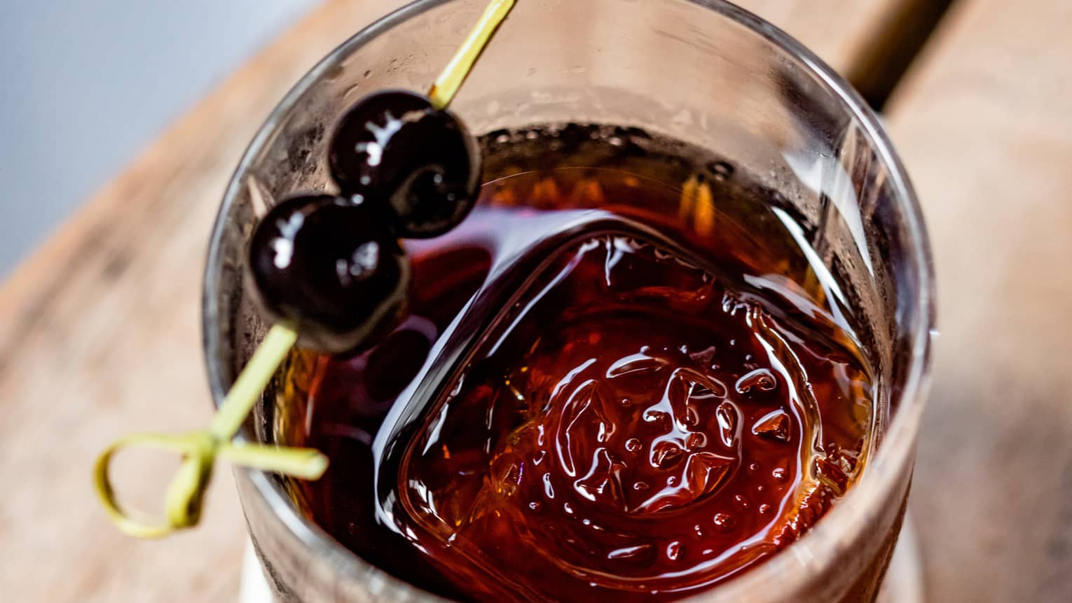 A dark coloured cocktail with two cherries on top of the glass.