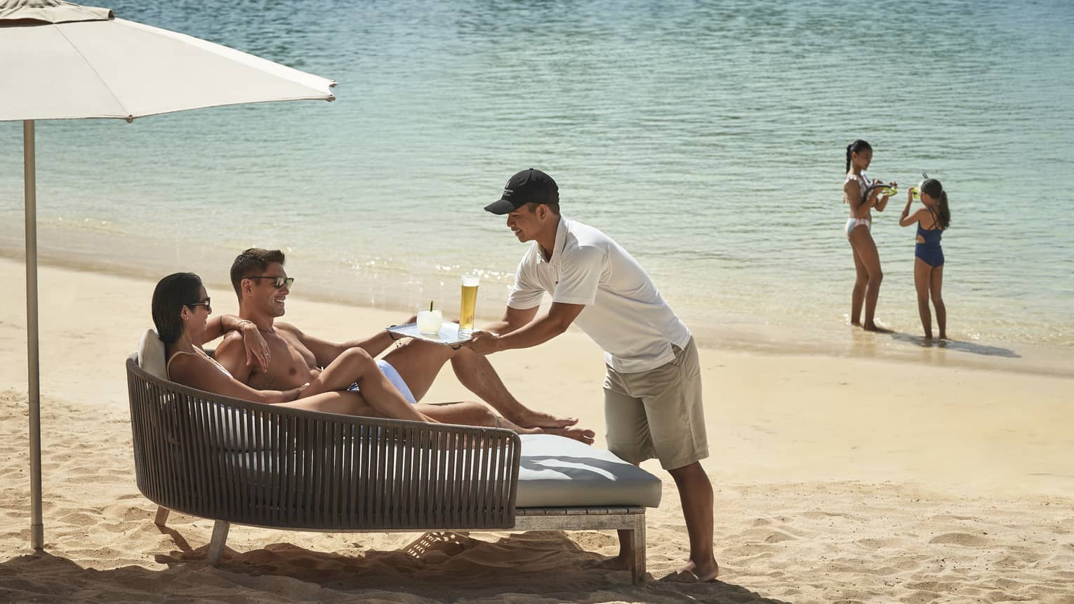 A man and woman get served a cocktail while lounging on daybed on a tropical beach