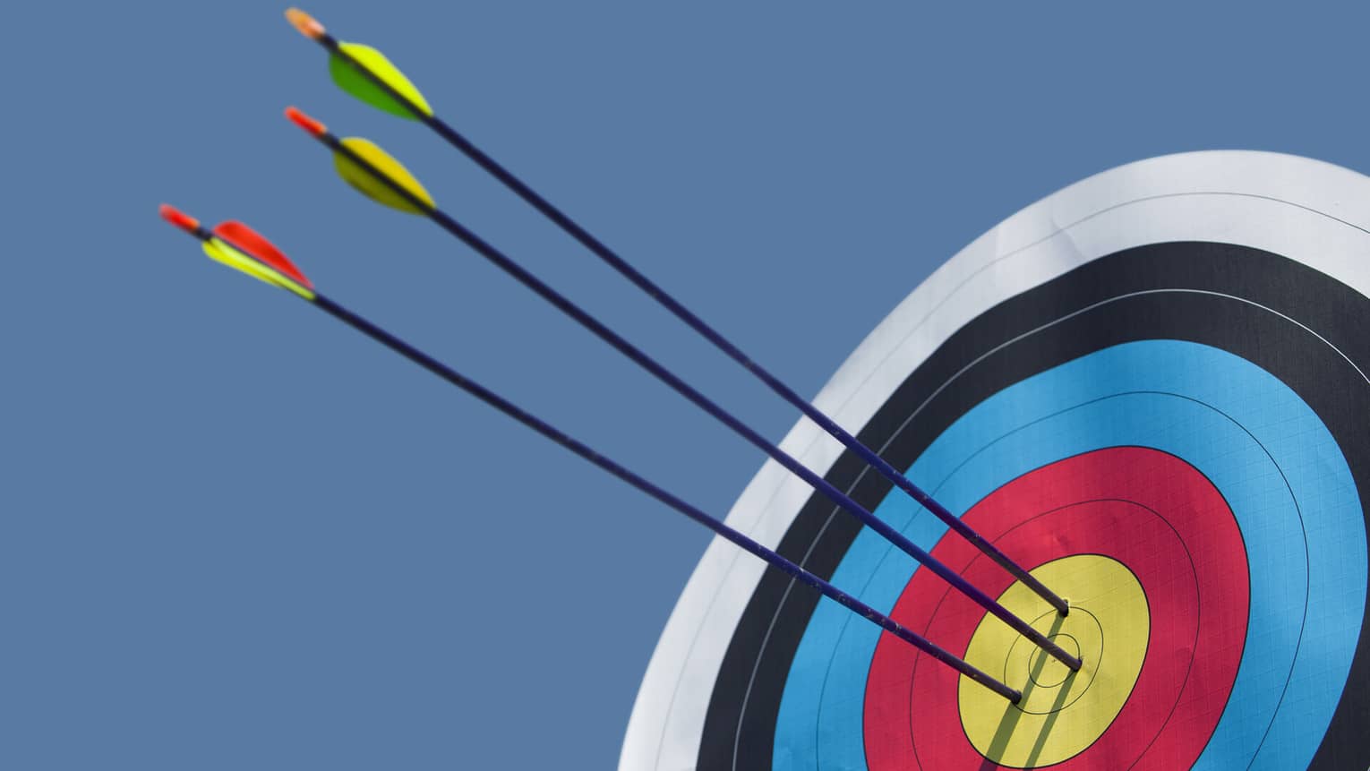 Three arrows with long black shafts pierce the gold circle at the centre of a bull's-eye against the backdrop of a blue sky.