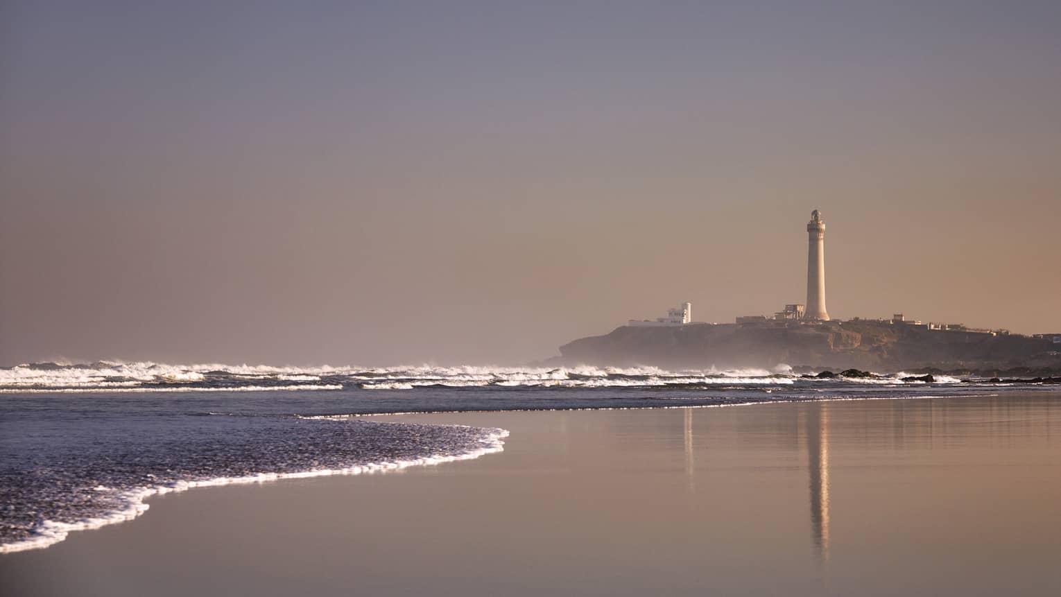 Flat seaside beach at sunrise with waves rolling in and El Hank Lighthouse in the distance