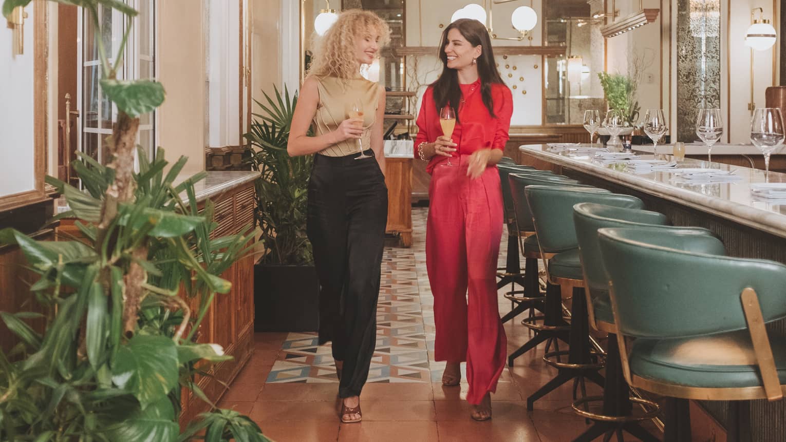 Two women walking a well lit restaurant with colourful tiles and seat at a bar.
