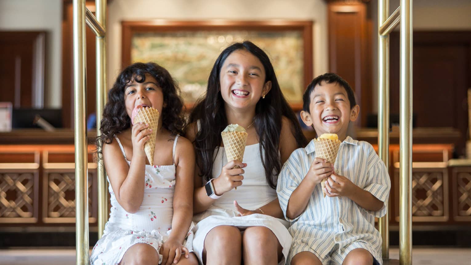 Three children sit on a gold hotel luggage cart eating ice cream