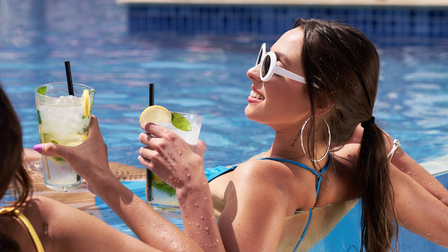 Person in swimsuit and sunglasses lounges in infinity-edge pool with drink in hand