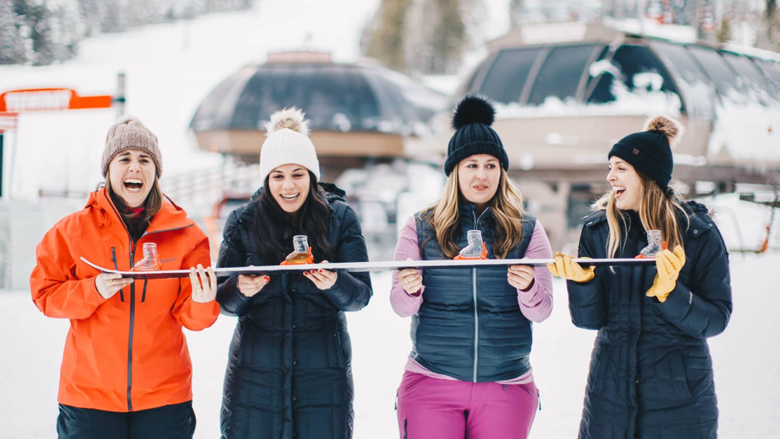 Four people in winter outdoor clothing stand in the snow, holding a ski, with snow-covered mountains in the background