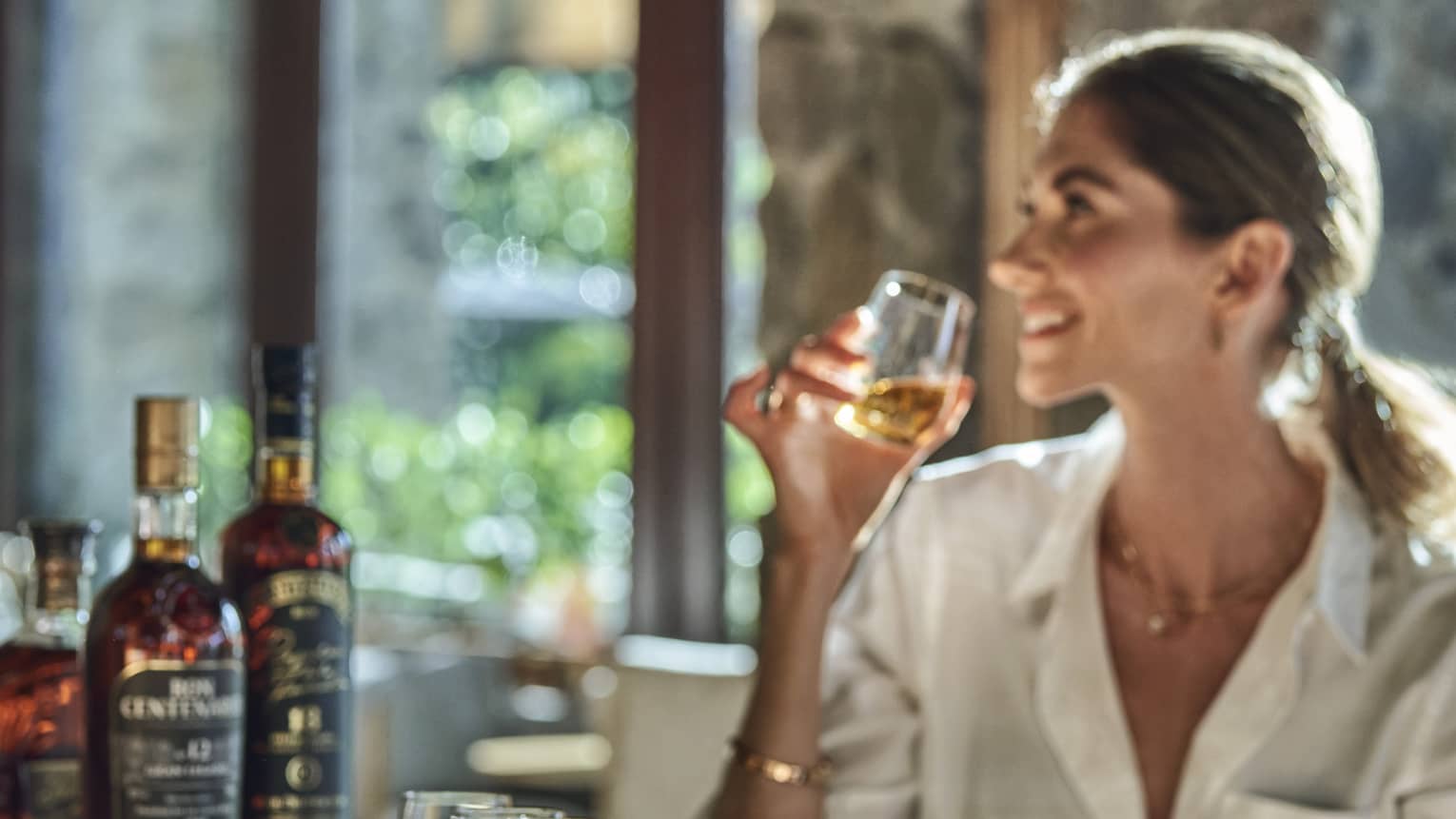 A guest smiles while holding a glass from a flight of eight rums, sun streaming in from behind through a bank of windows.