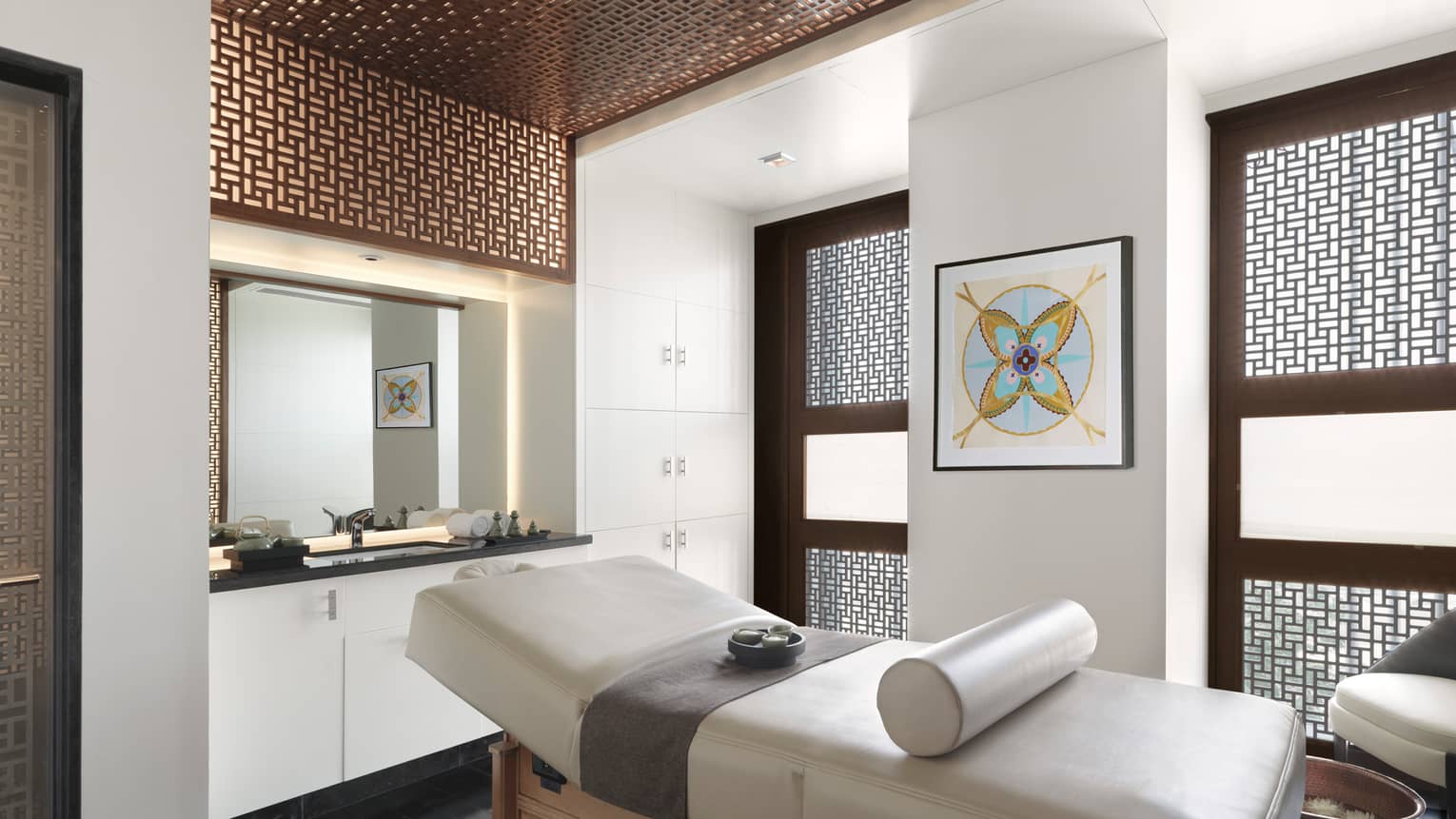 Spa massage table with rolled white leather pillow in front of vanity with sink, two windows