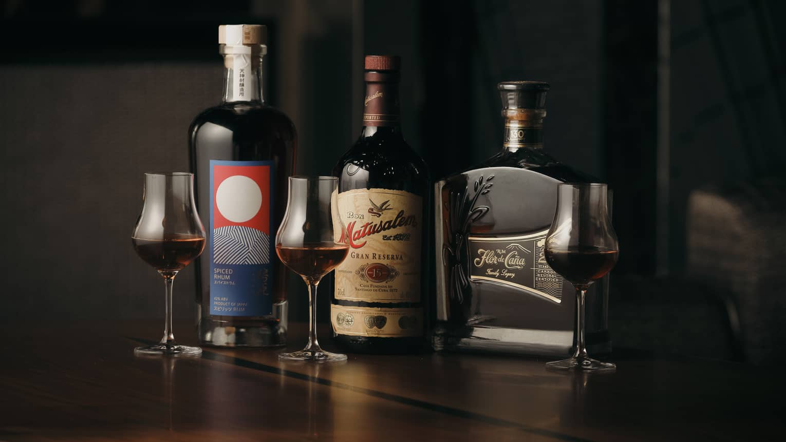 Dramatic dark lighting showcases three bottles of rum alongside three stemmed tulip-shaped glasses filled with dark rum.