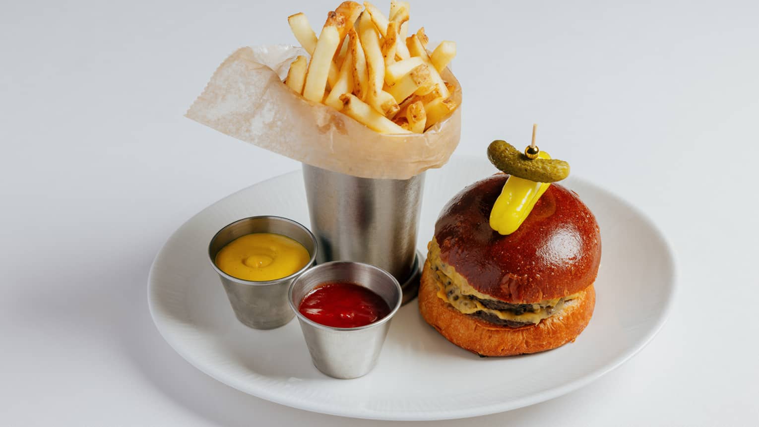 Burger topped with a yellow pepper and gherkin pickle on a toothpick plated next to a silver cup filled with French fries and small tins of ketchup and mustard