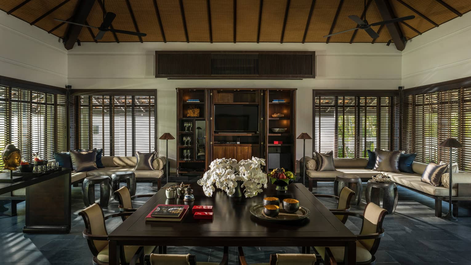 Large dining table with flowers in middle of room lined with sofas, windows, shutters