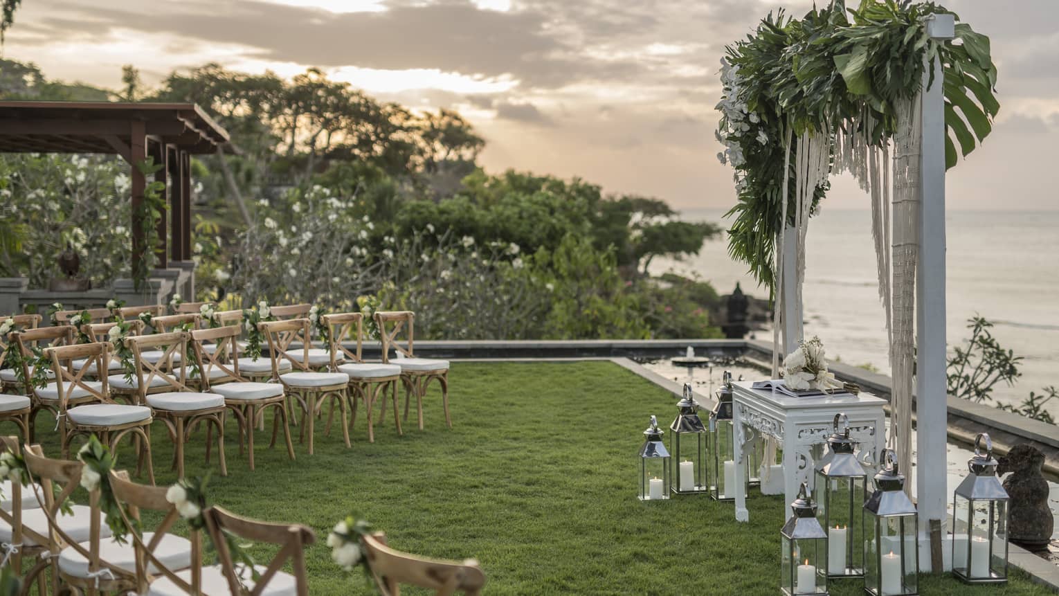 Rows of chairs face wedding altar with flowers, lanterns overlooking ocean at sunset