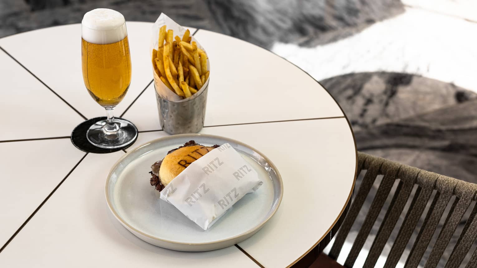 A burger on a plate next to a golden drink with foam on top in a clear stemmed glass and a container of French fries, all on a white table