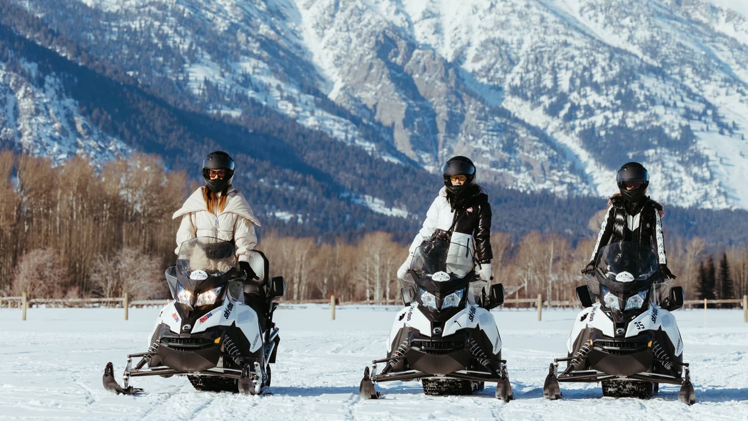 Three snowmobilers in stylish gear stand side-by-side on their machines facing the camera; a big snowy mountain looms behind.