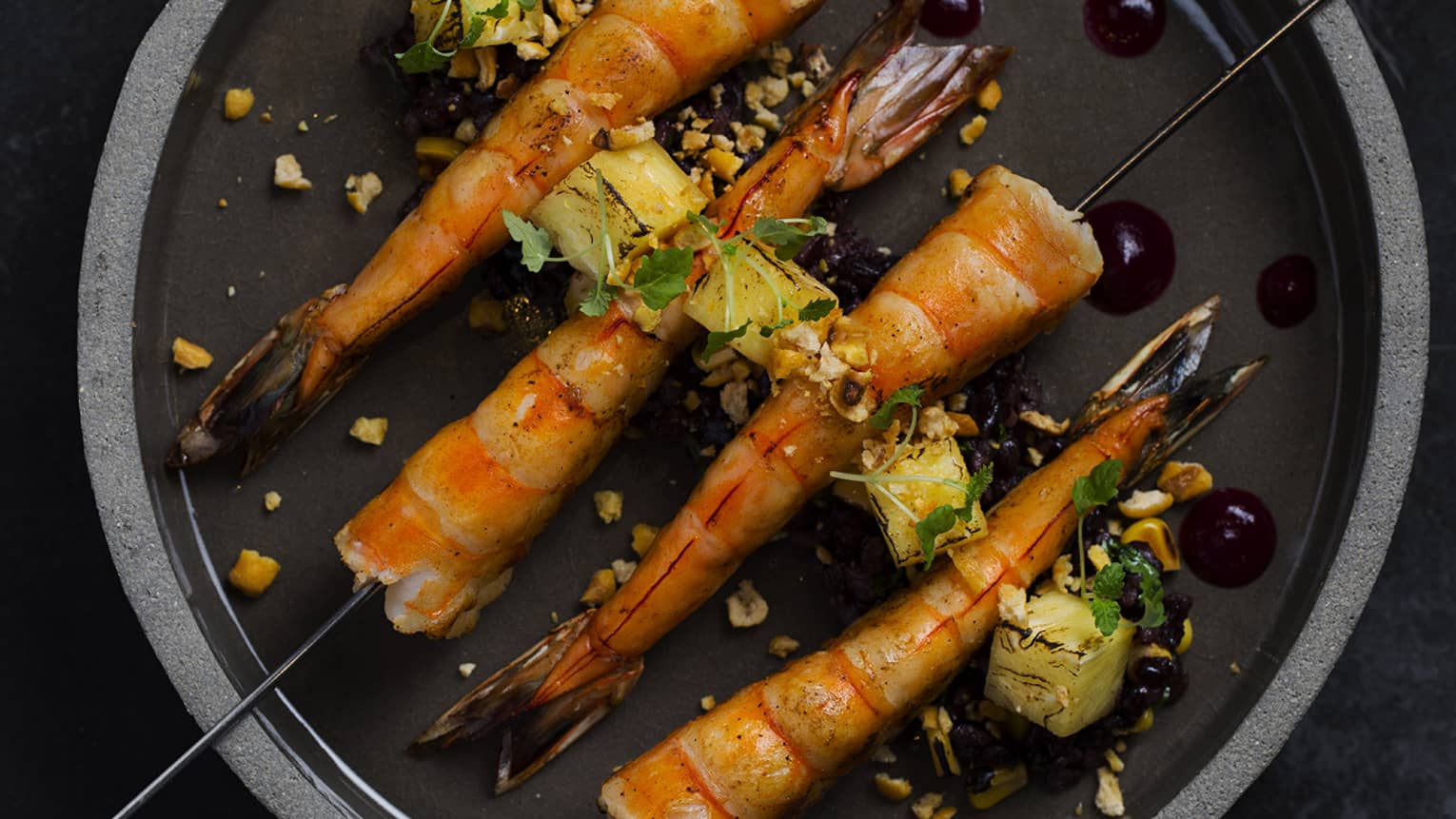 A stone bowl of Jumbo Prawns on metal skewers garnished with a variety of seeds