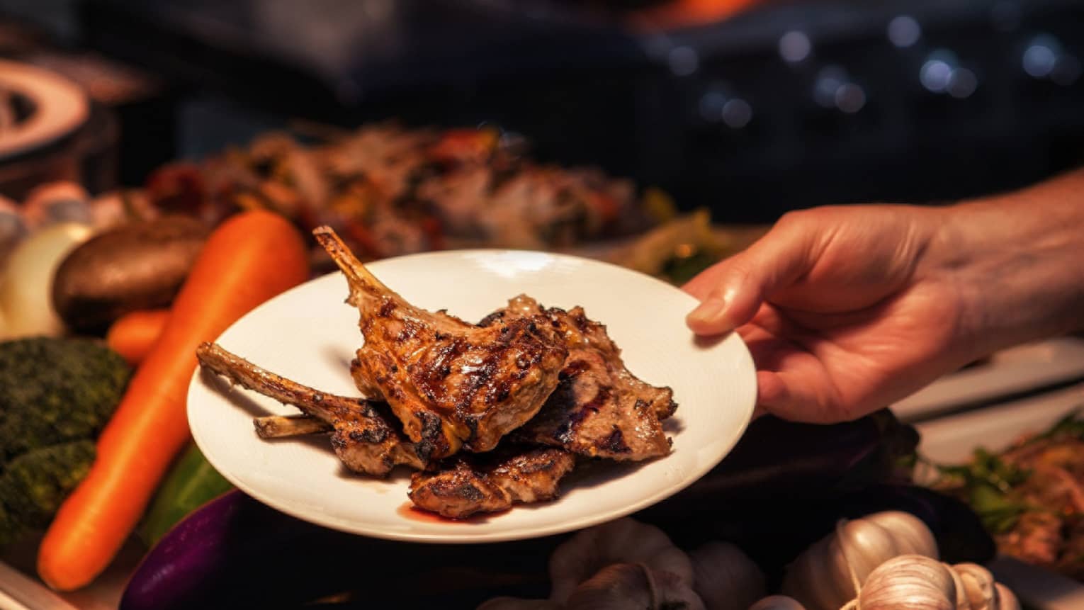 Hand holding barbecued chops on plate over fresh vegetables
