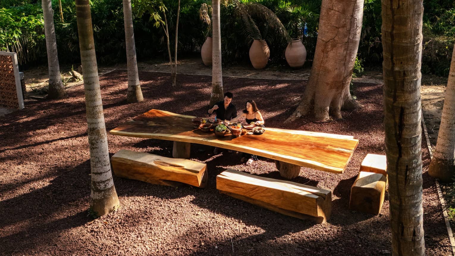 Two people sit at a long wood outdoor table with wood beam bench seats, all surrounded by palm trees.