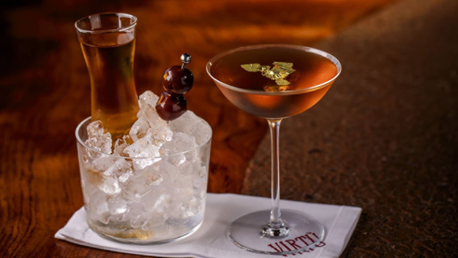 Two amber-coloured cocktails in stemmed glasses, one on ice with skewer of olives