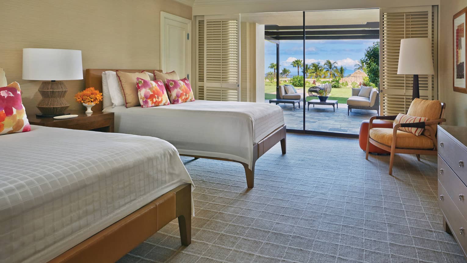 Presidential suite with two beds, colorful pillows and a seating area, leading to a patio with a garden and ocean view