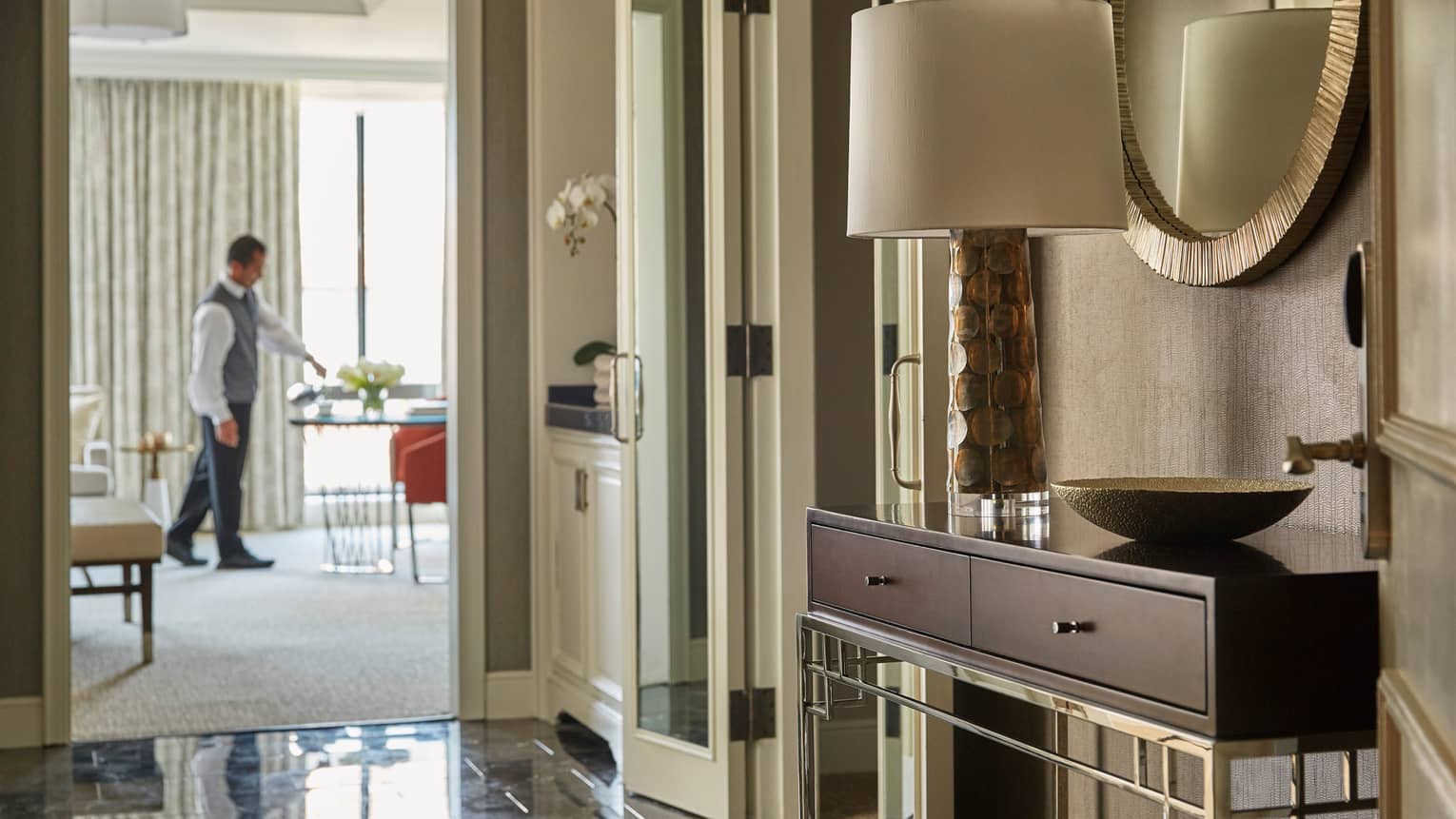 View from hotel suite hall with console table, lamp past doors to staff setting in-room dining table