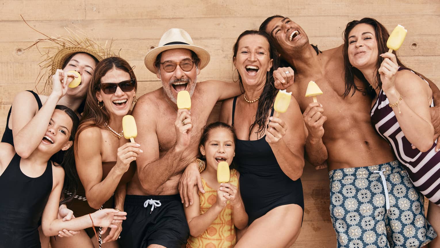 A group of adults and children in bathing suits hold yellow popsicles and smile at the camera.