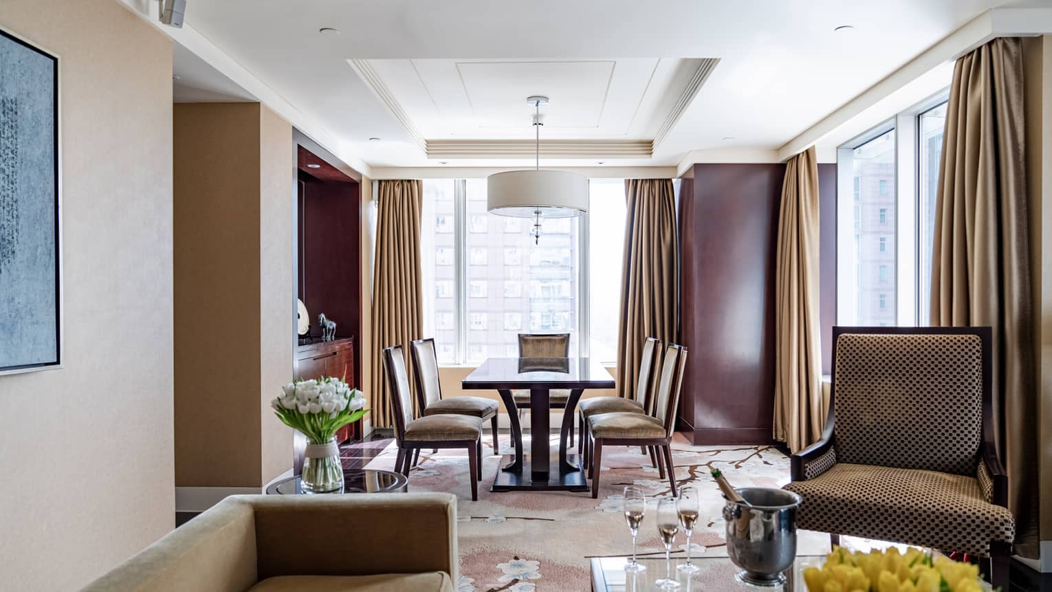 Tan couch and accent chair, glass coffee table, wooden dining table with four chairs, floor-to-ceiling windows