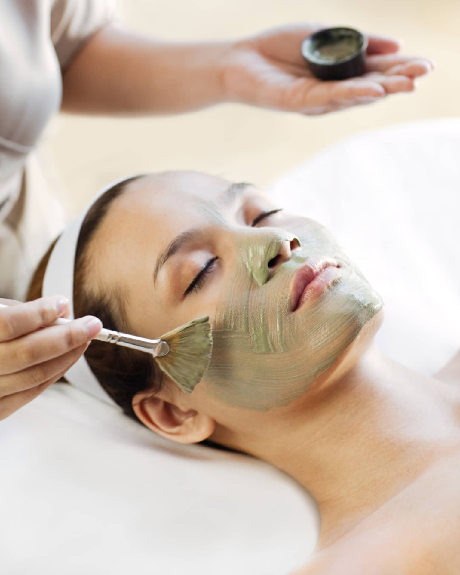 Spa staff holding brush paints green mask on woman's cheeks, chin, nose 