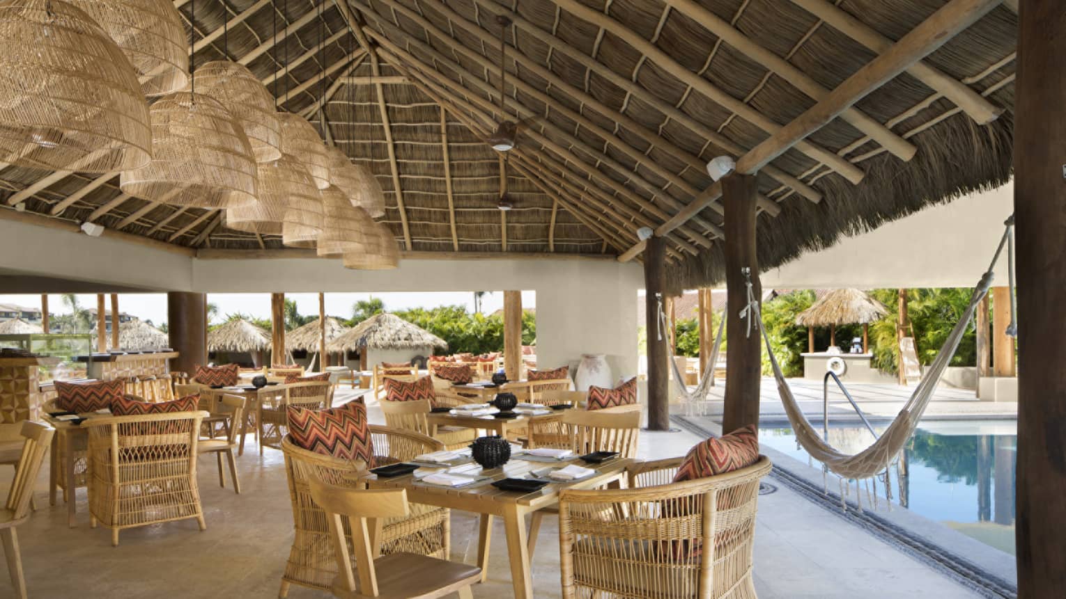 Whicker chairs and dining tables under thatched-roof pavilion, next to pool
