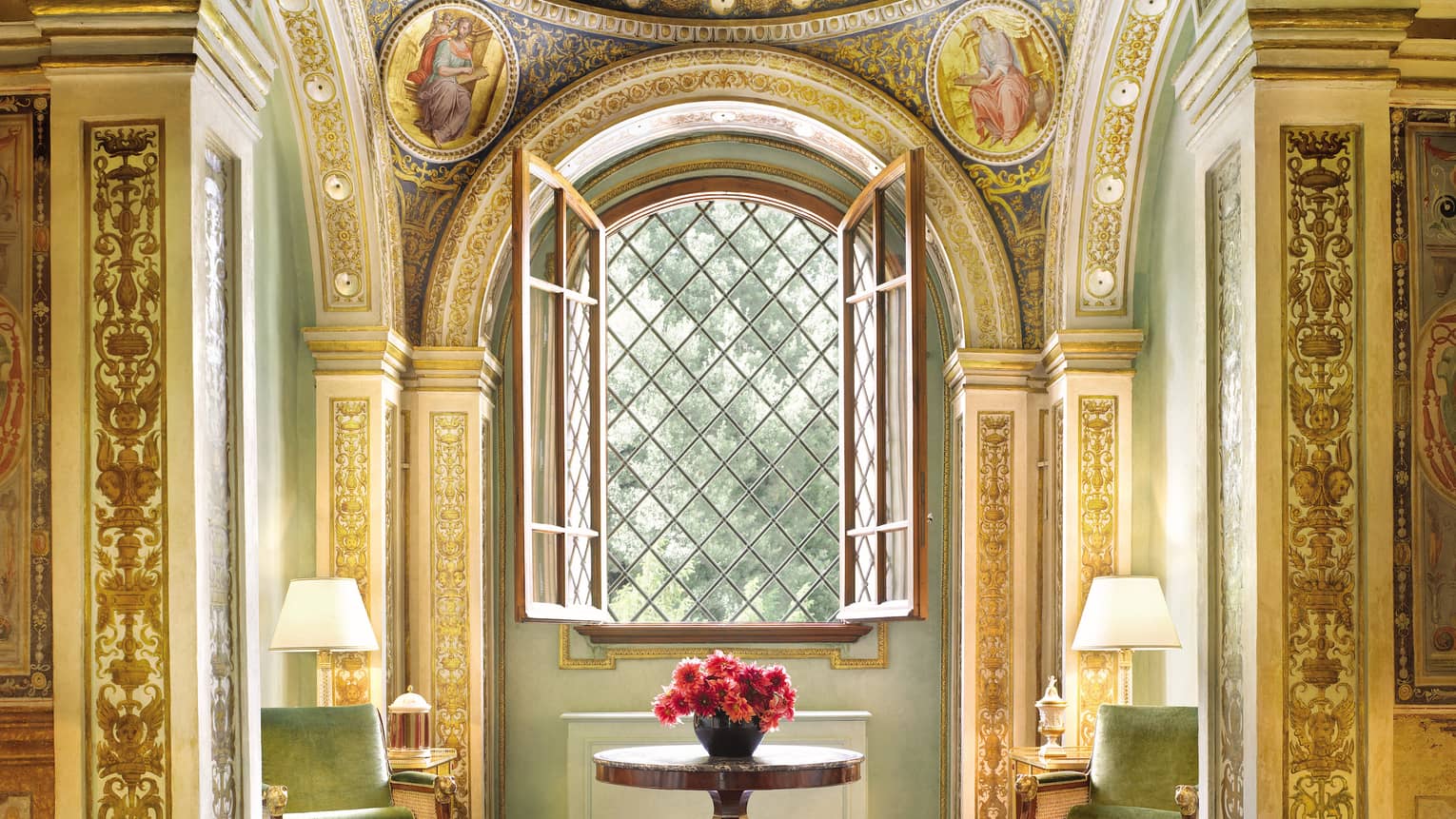 Green velvet armchairs around wood table with flowers, open window, gold ornate pillars