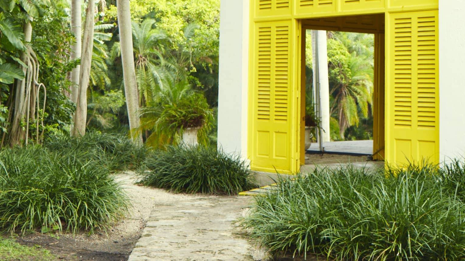 A cabana house with yellow shutter doors and a covered upper balcony with filigree railings is nestled amid lush foliage. 