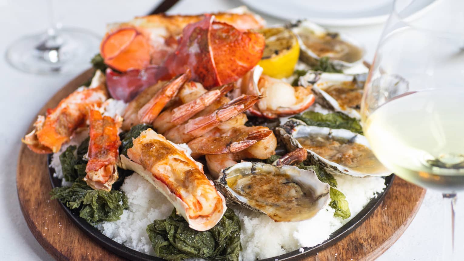 A wooden tray of seafood including oysters and crab, two glasses of wine are next to the tray.
