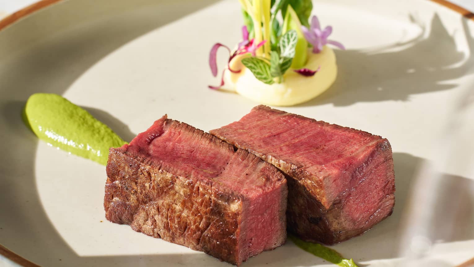 Two chunks of rare beef tenderloin with green sauce and a creamy dollop decorated with intricate leaves and petals.